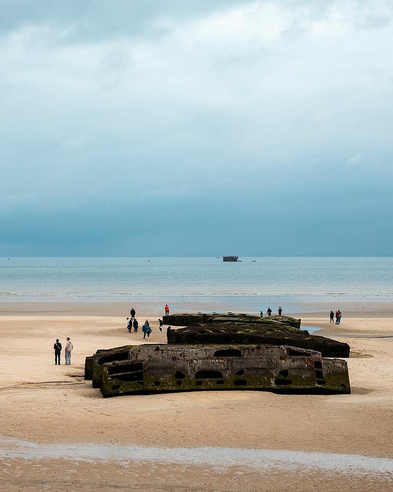 Wide shot of a sandy beach under an overcast sky, where tourists walk around large concrete blocks, an excursion easily accessible from Les Castels campings in Normandy.