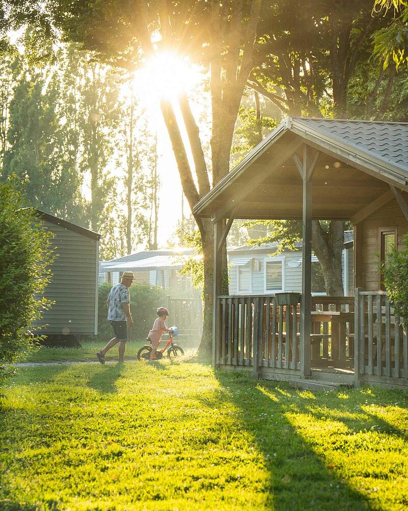 A grandfather walking alongside a young child riding a bike at sunset between wooden chalets in a wooded campsite near Saint-Malo.