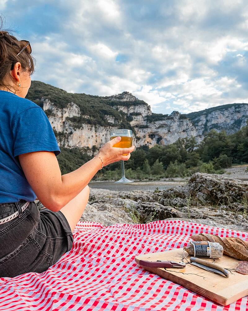 Woman enjoying a drink during a picnic on a red checkered blanket by the river in front of high rock cliffs in Ardèche with Les Castels.