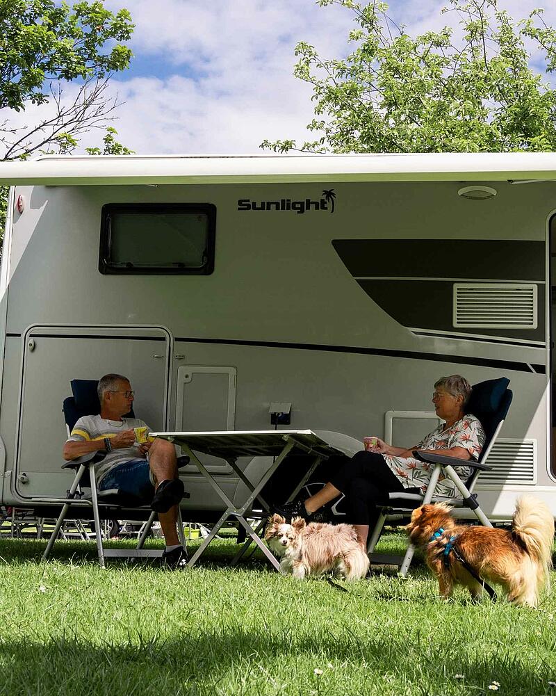 A man and a woman sitting at a camping table under the awning of a modern Sunlight motorhome on a pet-friendly pitch at Les Castels.