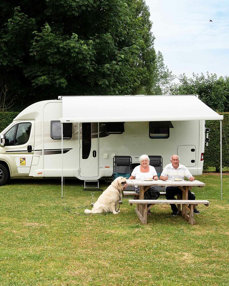 A senior couple enjoying a meal at a wooden picnic table outside their white motorhome under an awning, accompanied by their golden retriever dog on a green campsite pitch.