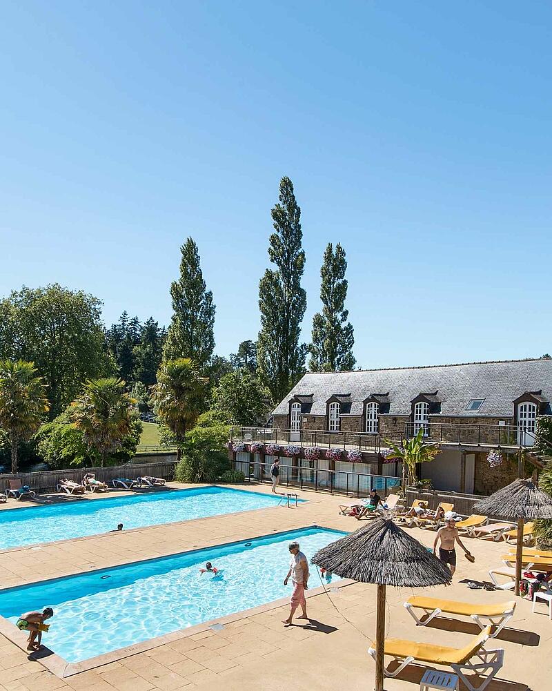Large outdoor swimming pool surrounded by sun loungers, straw parasols, and palm trees, in front of a traditional stone building at Domaine des Ormes in Dol-de-Bretagne.