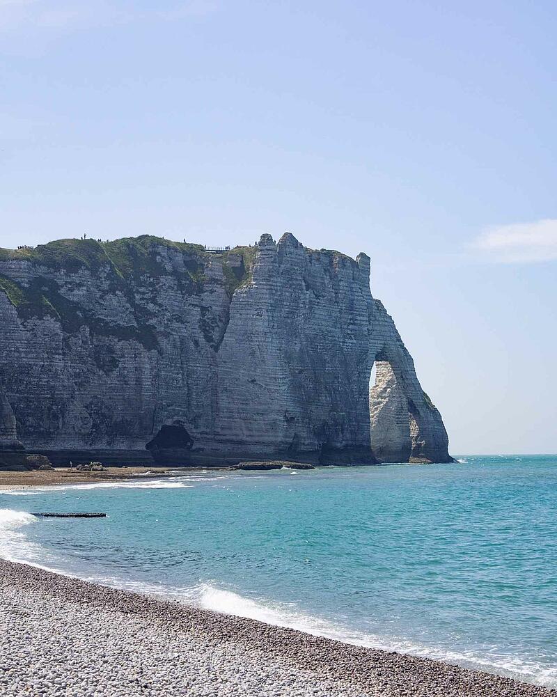 The dramatic white chalk cliffs and natural rock arch of Étretat rising above a pebble beach and the turquoise waters of the sea in Normandy.