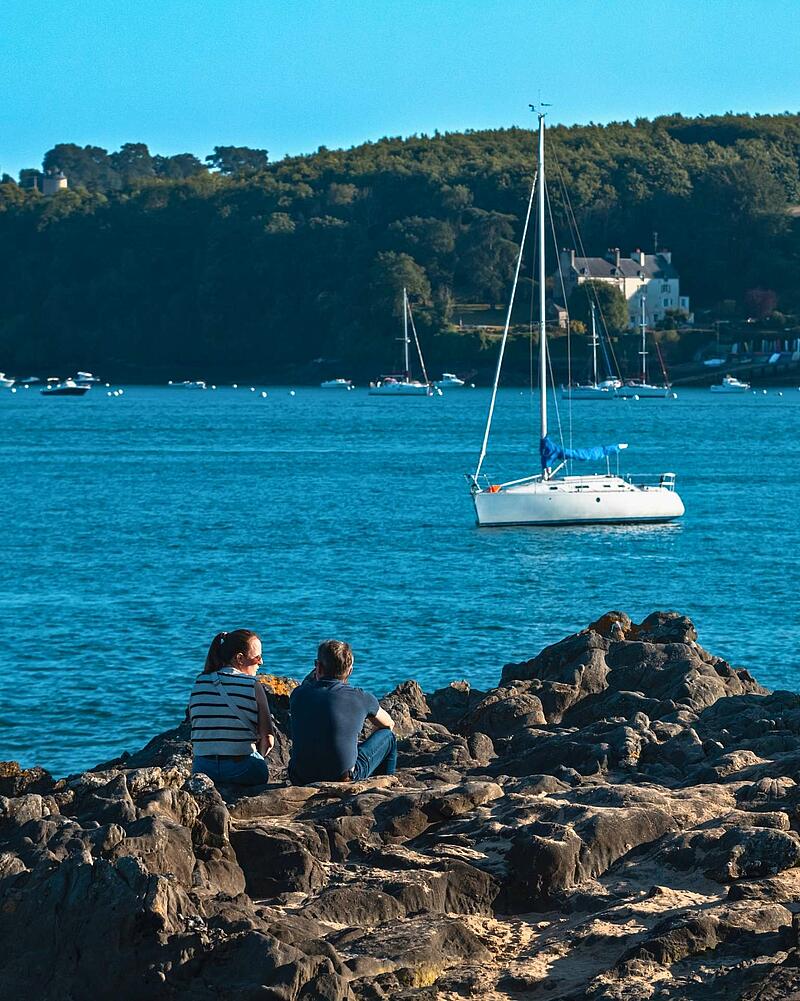 A couple sit relaxing on rugged, dark rocks overlooking a tranquil bay filled with moored sailboats and a treed coastline in the distance, capturing the peaceful atmosphere near Les Castels seaside campings in France.