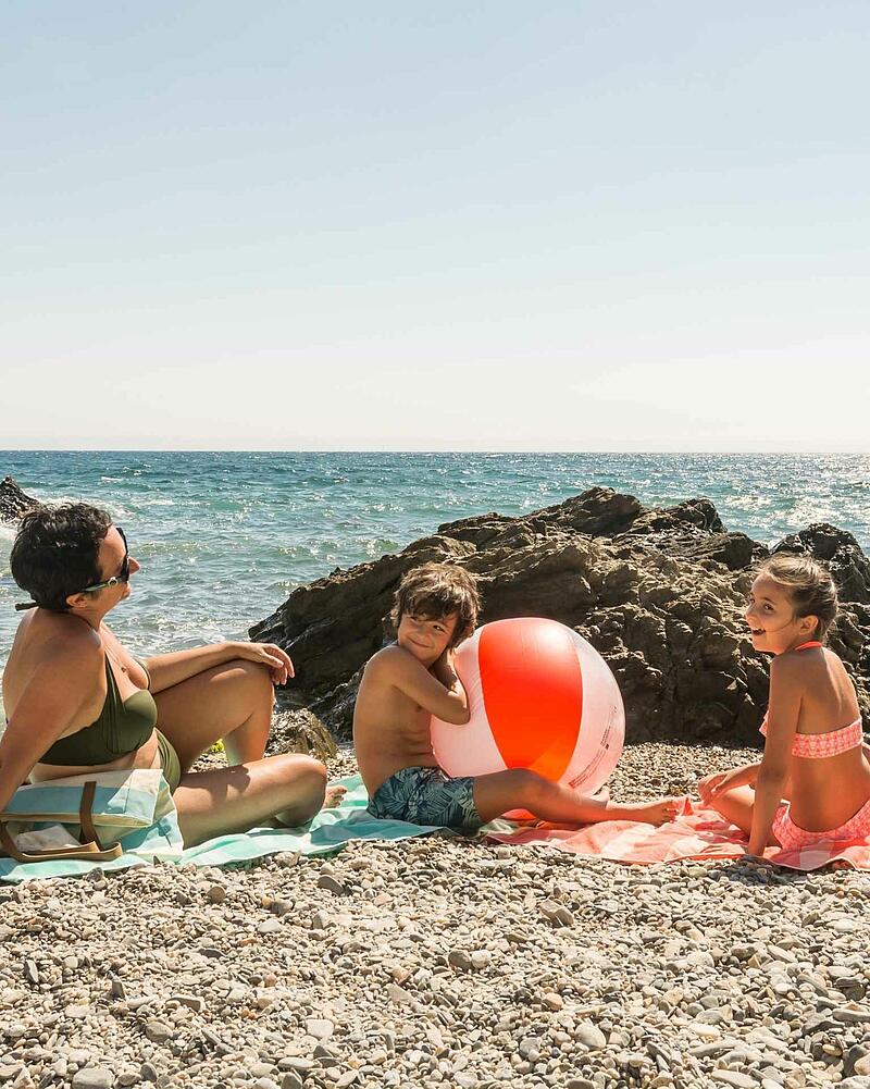 A mother and two children sitting on a pebble beach with a large orange beach ball, enjoying a sunny family holiday at a French seaside campsite