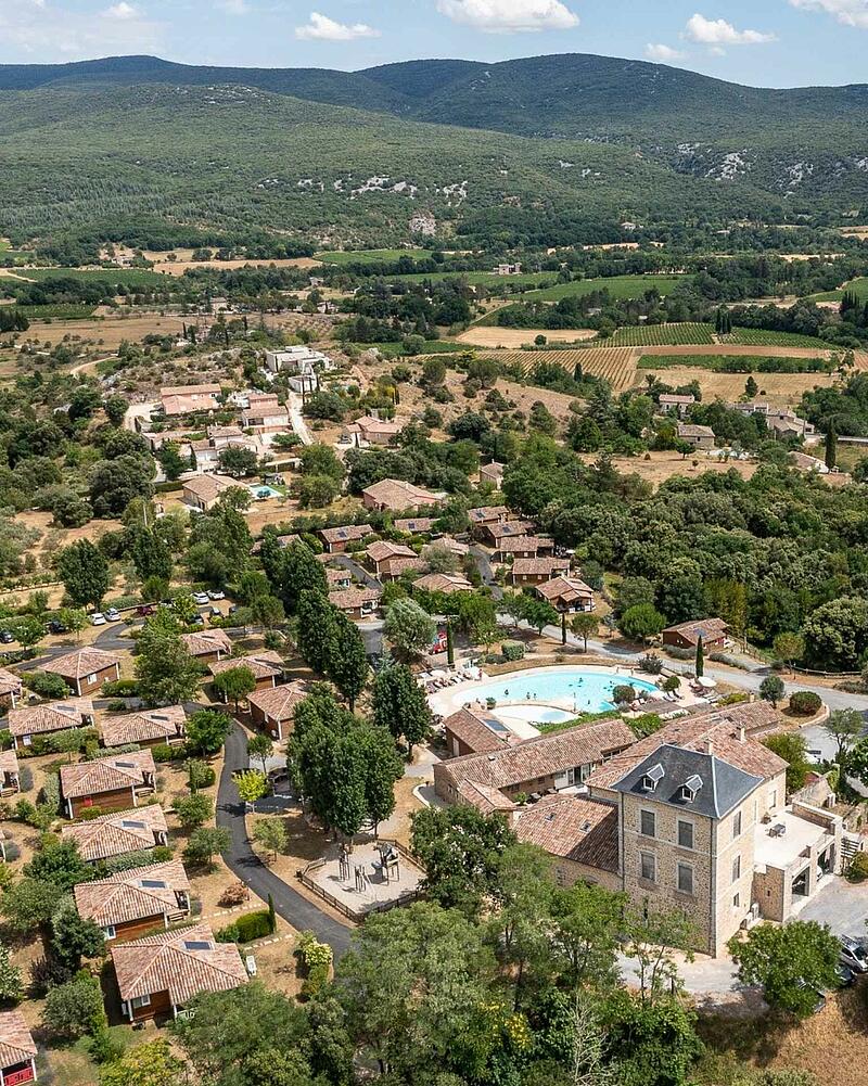 Drone perspective of the heated outdoor swimming pool, stone estate building, and eco-friendly wooden chalets surrounded by nature at a 5-star Les Castels campsite.