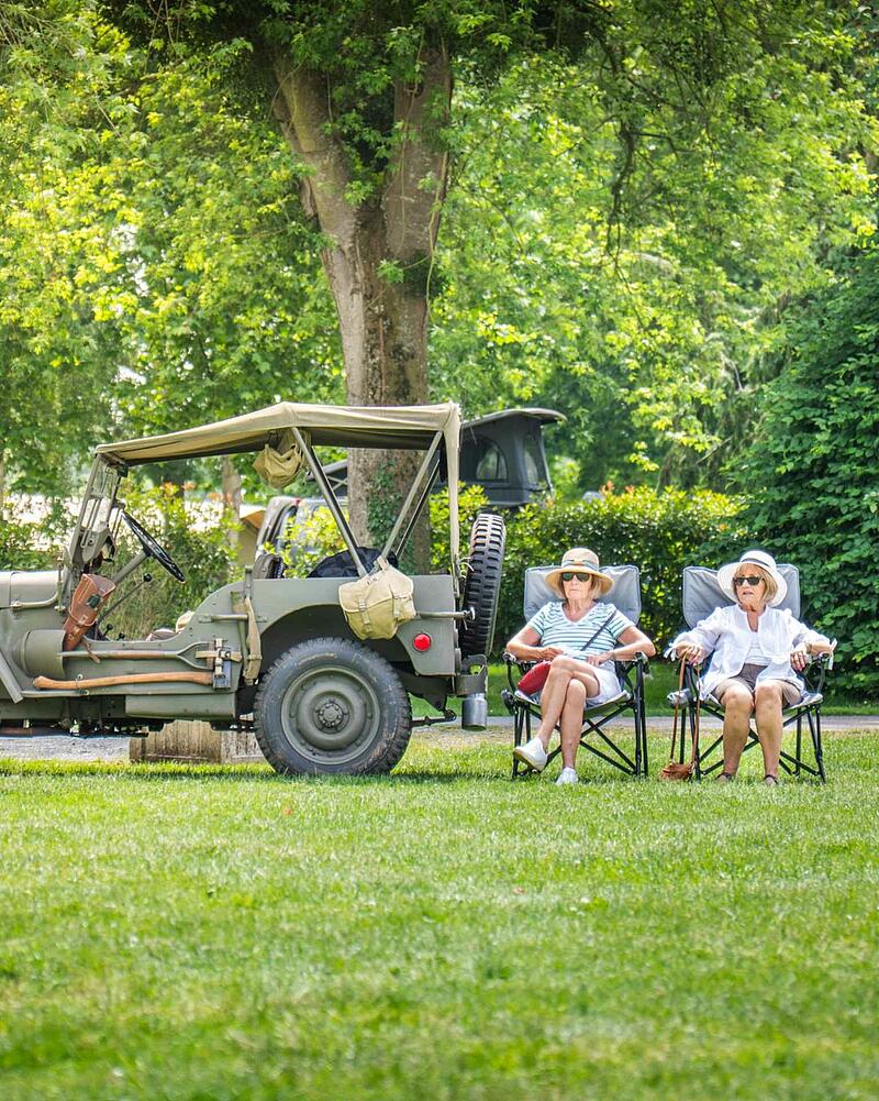 Two senior women wearing sun hats sitting on camping chairs on a green lawn next to a classic green vintage military jeep at a Les Castels campsite.