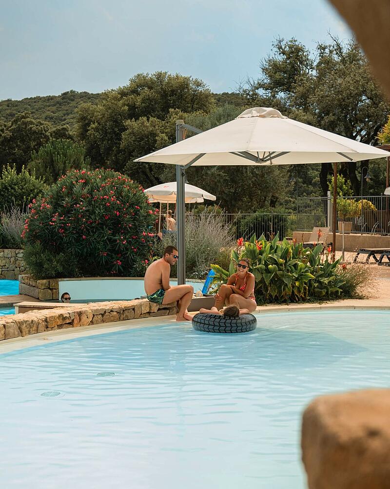Couple relaxing by a sunlit swimming pool area with large umbrellas during a last-minute stay at Les Castels luxury campsites in Ardèche.