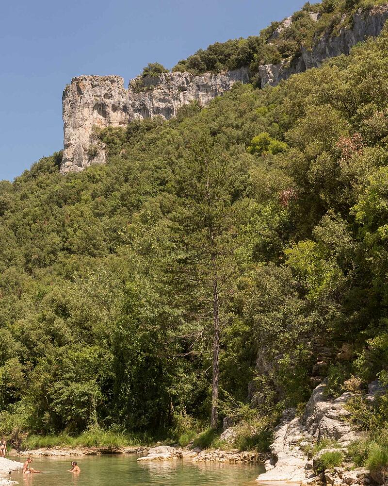 People swimming and relaxing in the clear river water at the foot of towering limestone cliffs in the Ardeche Gorges near a luxury Les Castels estate.