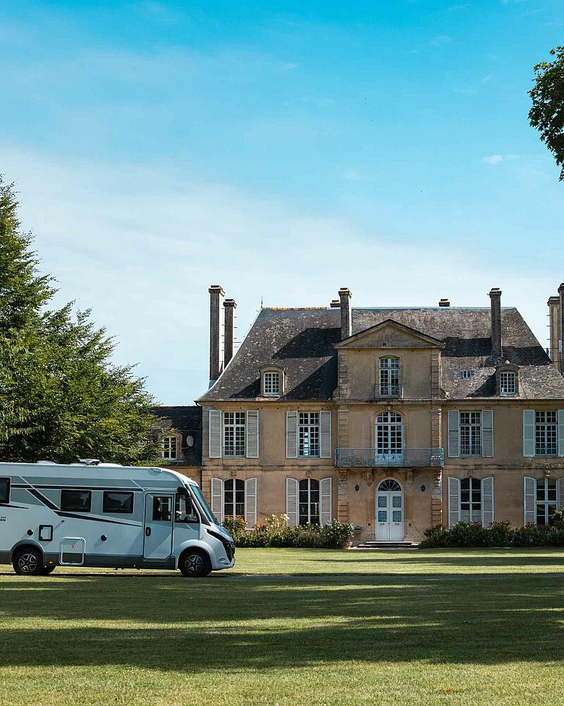 A sleek white motorhome parked on the manicured lawns in front of a grand, historic French manor house during a summer road trip.