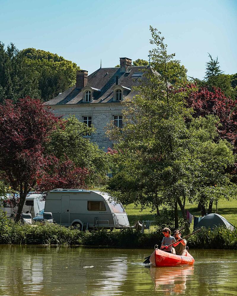 Two people canoeing on a lake in the foreground, with a caravan parked on the shore and a grand traditional French chateau partially hidden by trees in the background at Les Castels authentic 5-star campsites in France, illustrating premium campsites France.