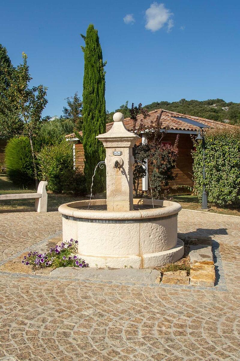 Fontaine ancienne en pierre avec chalets écologiques en arrière-plan, illustrant le charme du patrimoine français dans un domaine de plein air.