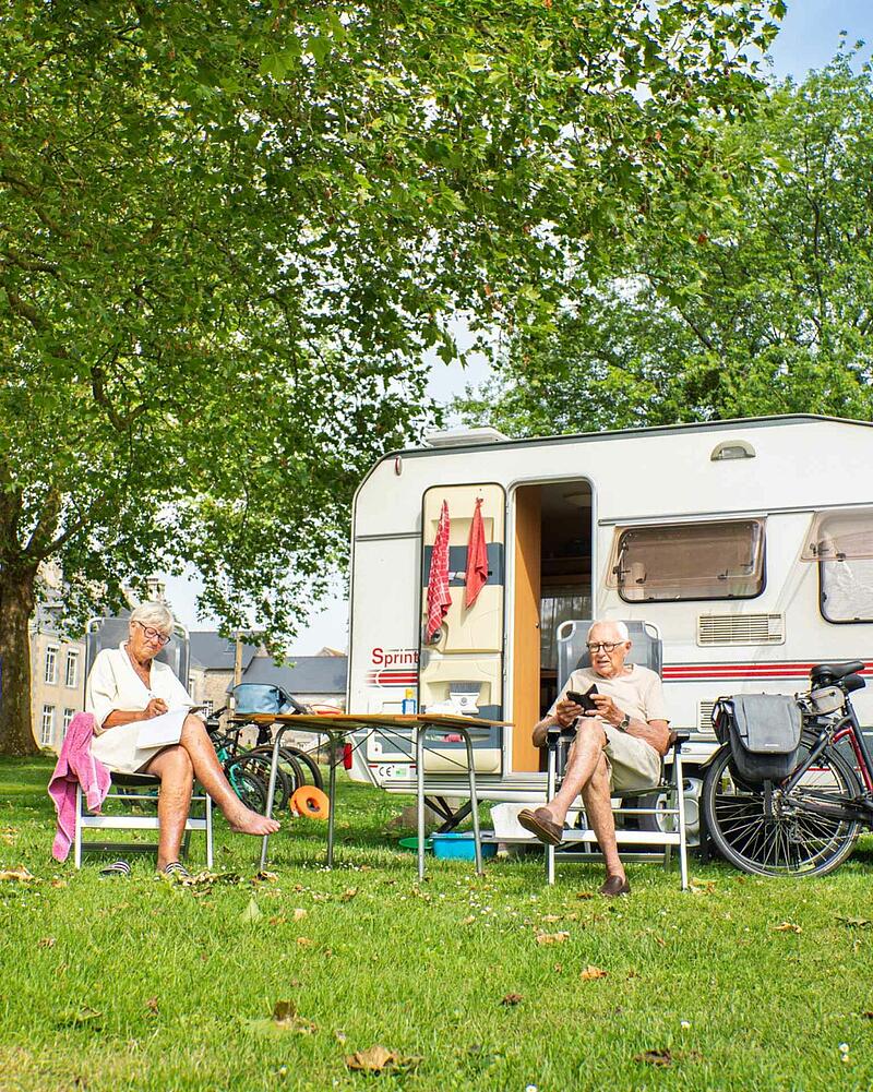 An elderly couple relaxes in camping chairs next to their white caravan on a lush lawn, with a bicycle parked nearby and a historic stone building in the background.