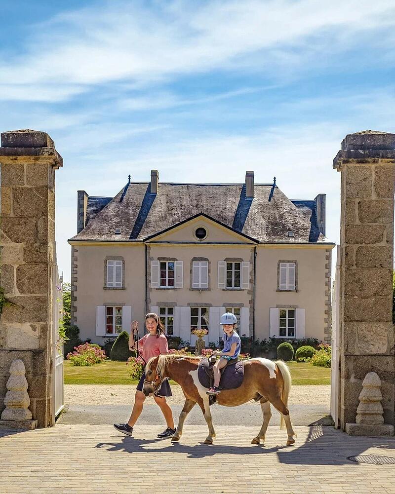 A child wearing a riding helmet sits atop a pony led by an instructor, walking past grand stone pillars in front of a majestic historic chateau at a Les Castels campsite.