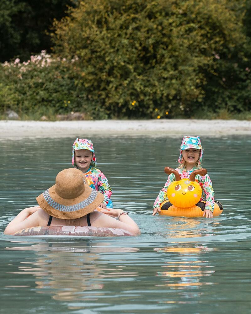 Two smiling young children wearing sun-protection swimwear float in the clear turquoise water of a premium Les Castels natural lagoon, alongside an adult wearing a straw hat.