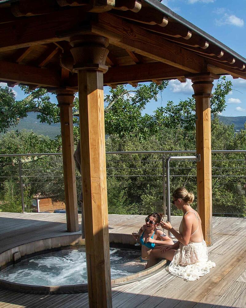 Two women enjoying drinks in an outdoor wooden hot tub with scenic countryside views at Les Castels 5-star estate campsite in Vallon-Pont-d'Arc.