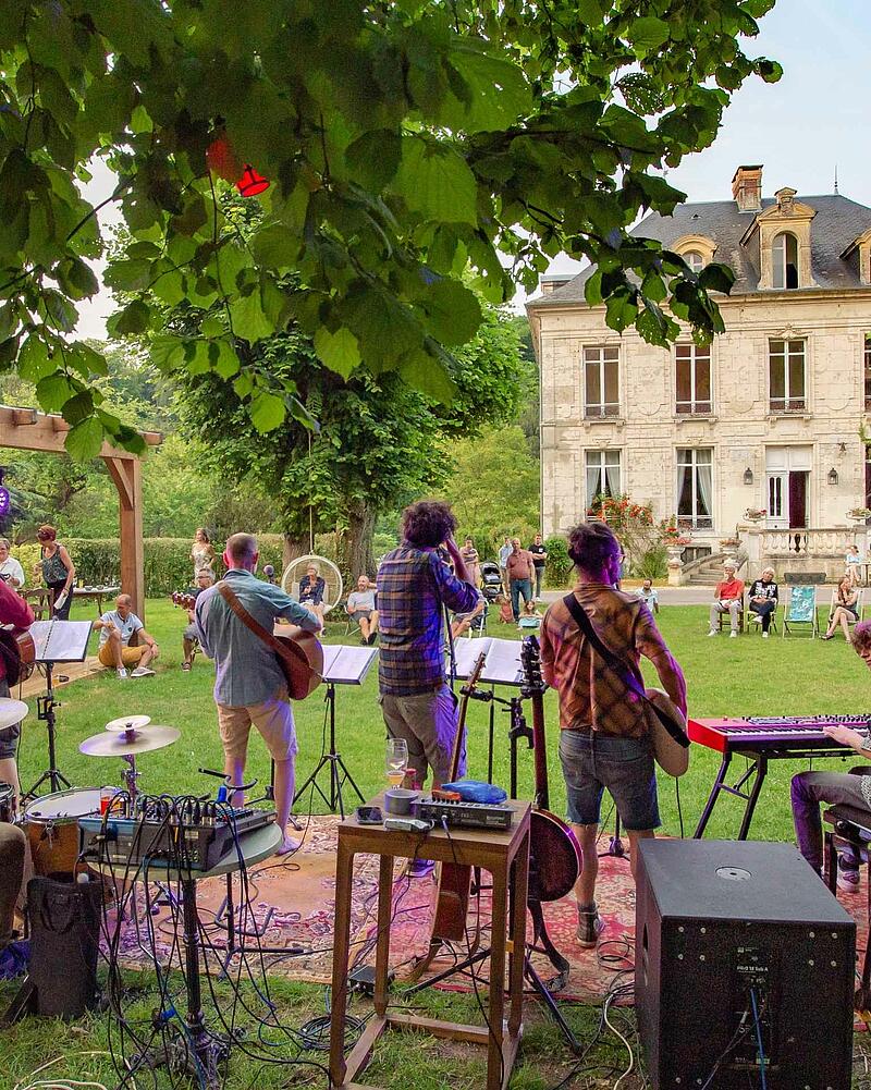 Stage view from behind a live band playing guitars and keyboards outdoors for an audience seated on the grass during a premium evening event at Les Castels.