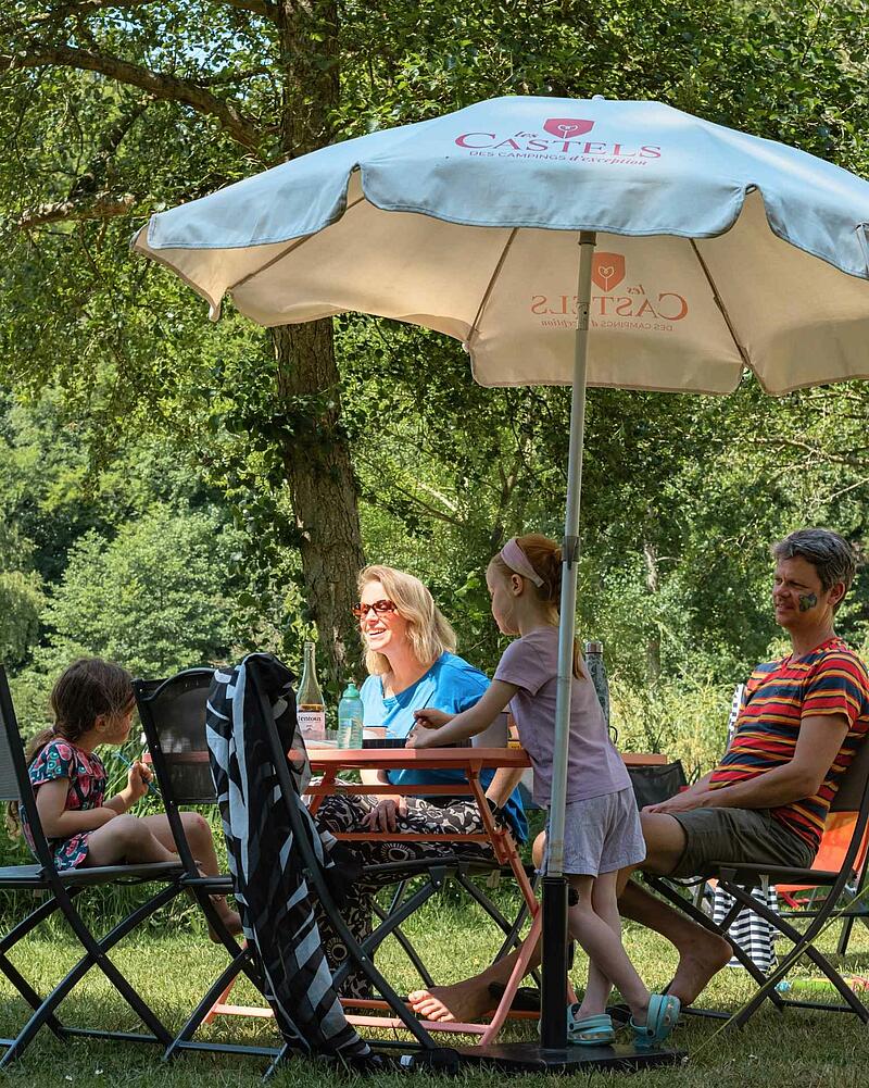 A cheerful family shares a joyful picnic under the shade of a tree, capturing the happy moments and friendly atmosphere to be found on a last-minute camping break at Les Castels