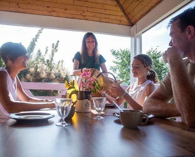 Family breakfast on the chalet terrace at Castels Domaine de Sévenier, Ardèche, France. A family gathers around a wooden table on a shaded chalet terrace at Castels camping Domaine de Sévenier.