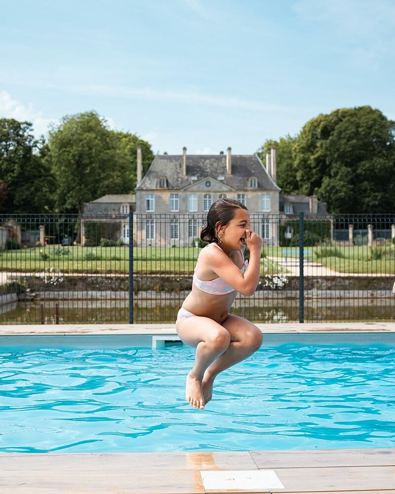 A young girl is captured mid-air, happily leaping into a large, crystal-clear swimming pool with a grand French castle in the background, illustrating the fun and premium pool facilities for an impromptu summer vacation at Les Castels,