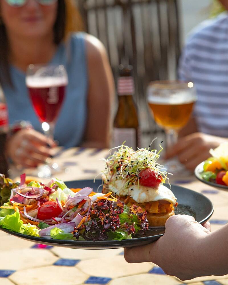 A waiter serving a fresh, colorful salad platter with sprouts and a fried egg on toast at an outdoor terrace, with campsite guests enjoying beer and wine in the blurred background.