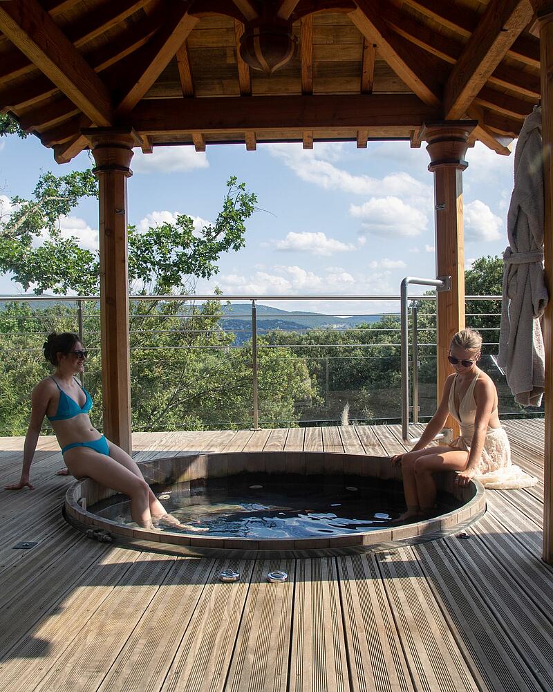 Two women relaxing in an outdoor wooden jacuzzi with a scenic forest view at Les Castels spa campsites in Ardèche.