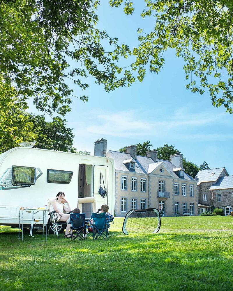 A white touring caravan parked on a wide grassy field next to a grand French chateau, with a family sitting outside under the trees.