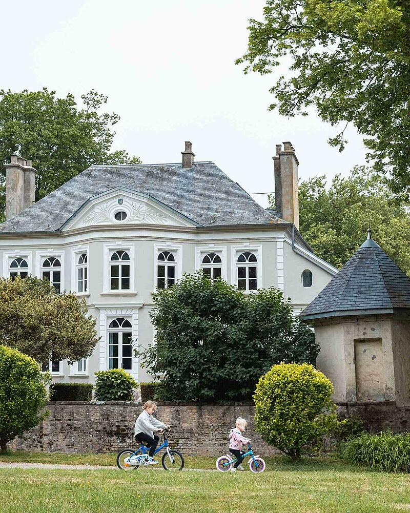 Children cycling in front of the historic manor house at Camping La Bien-Assise, a premium Les Castels stopover campsite located near Calais Port.