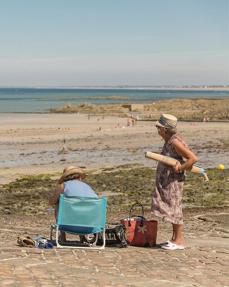 Two women wearing straw hats spending a sunny day on a stone seawall, overlooking a vast sandy beach during a seaside holiday with Les Castels campings.