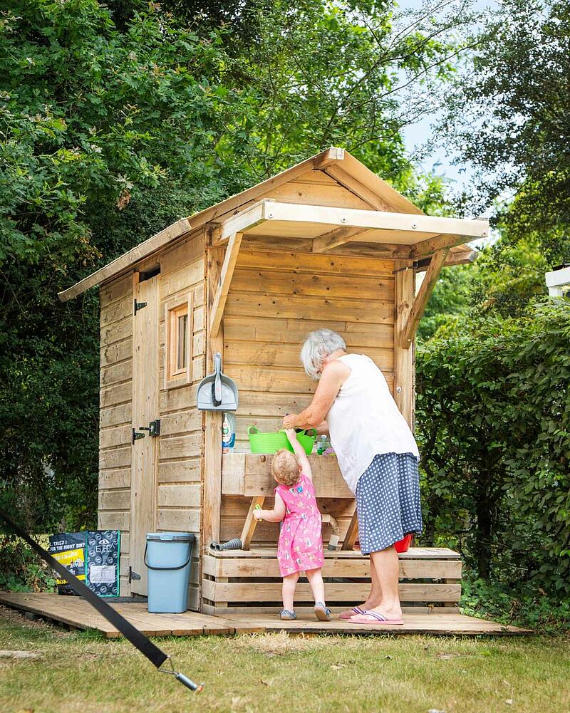 Woman and young girl interact at a rustic wooden outdoor kitchen station on a Les Castels camping estate.