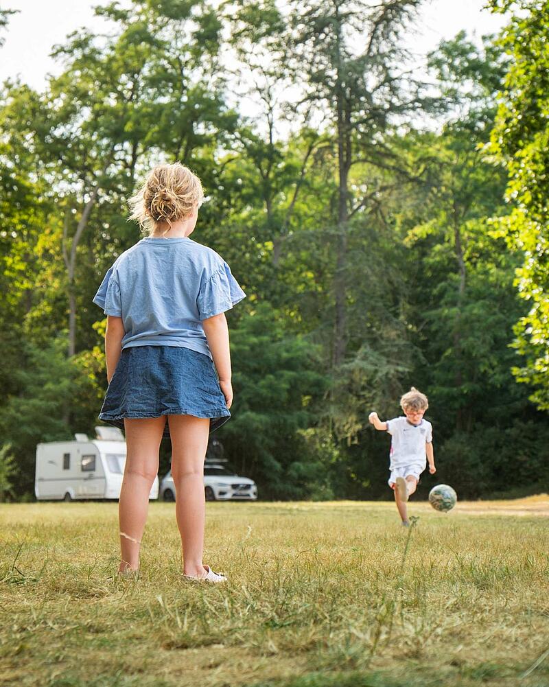 From behind, a girl in a denim skirt watches a boy in a football kit kick a ball on a large grassy field at a Les Castels site, with trees and a caravan in the distance.