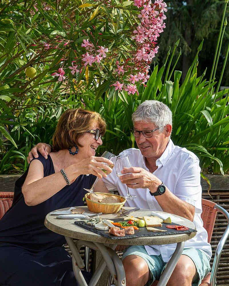 A man and a woman laughing and clinking wine glasses at an outdoor table, enjoying an artisanal cheese board at a premium Les Castels campsite restaurant.