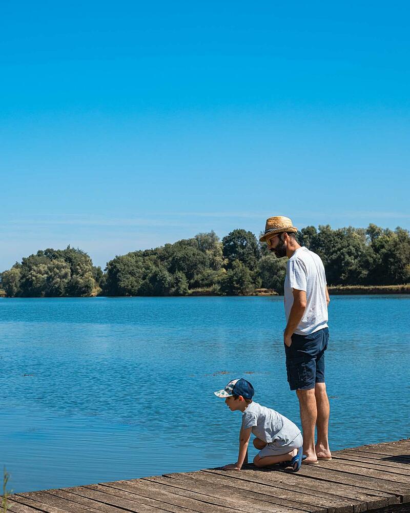 A father wearing a straw hat stands next to his young son who is looking into the calm blue water from a wooden jetty on a private campsite lake.