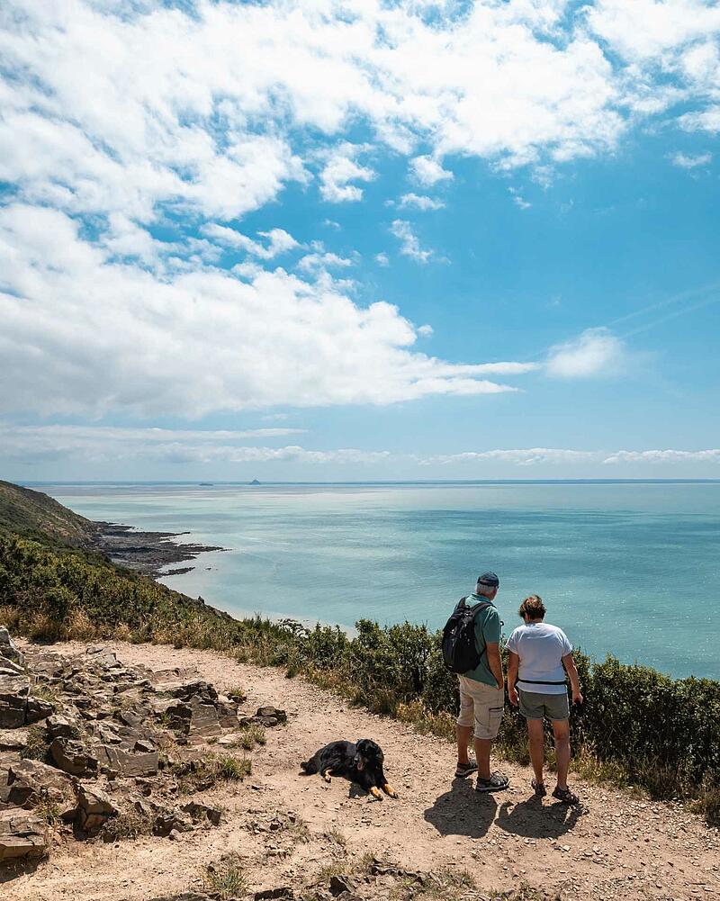 Two hikers and a dog standing on a cliffside trail overlooking the turquoise waters of Mont Saint-Michel Bay during a coastal camping trip in Normandy.