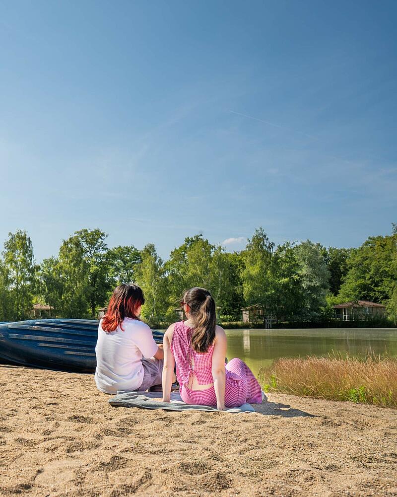 Two friends are relaxing on a sandy beach next to a tranquil lake with small rowboats, surrounded by trees, highlighting the restful lakeside chalet accommodations available for booking at the last minute at Les Castels