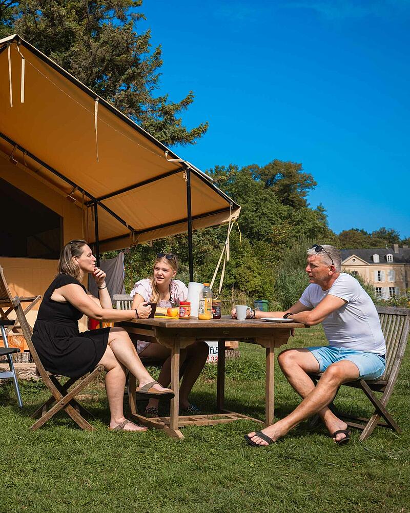 A mother, father, and daughter sit around a wooden picnic table having breakfast outside a large luxury canvas glamping safari tent on a sunny campsite lawn.