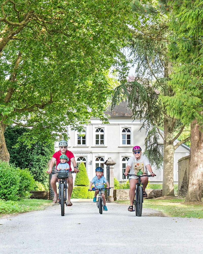 A family of four, including two young children wearing helmets, cycling towards the camera on a gravel path during their holiday at a Les Castels domain.