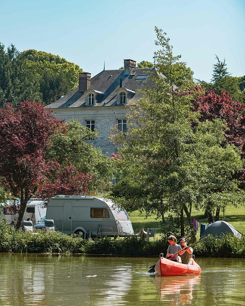 An orange canoe with a man and a young boy paddling on a calm lake, with a caravan parked on the grassy bank of a Les Castels estate.