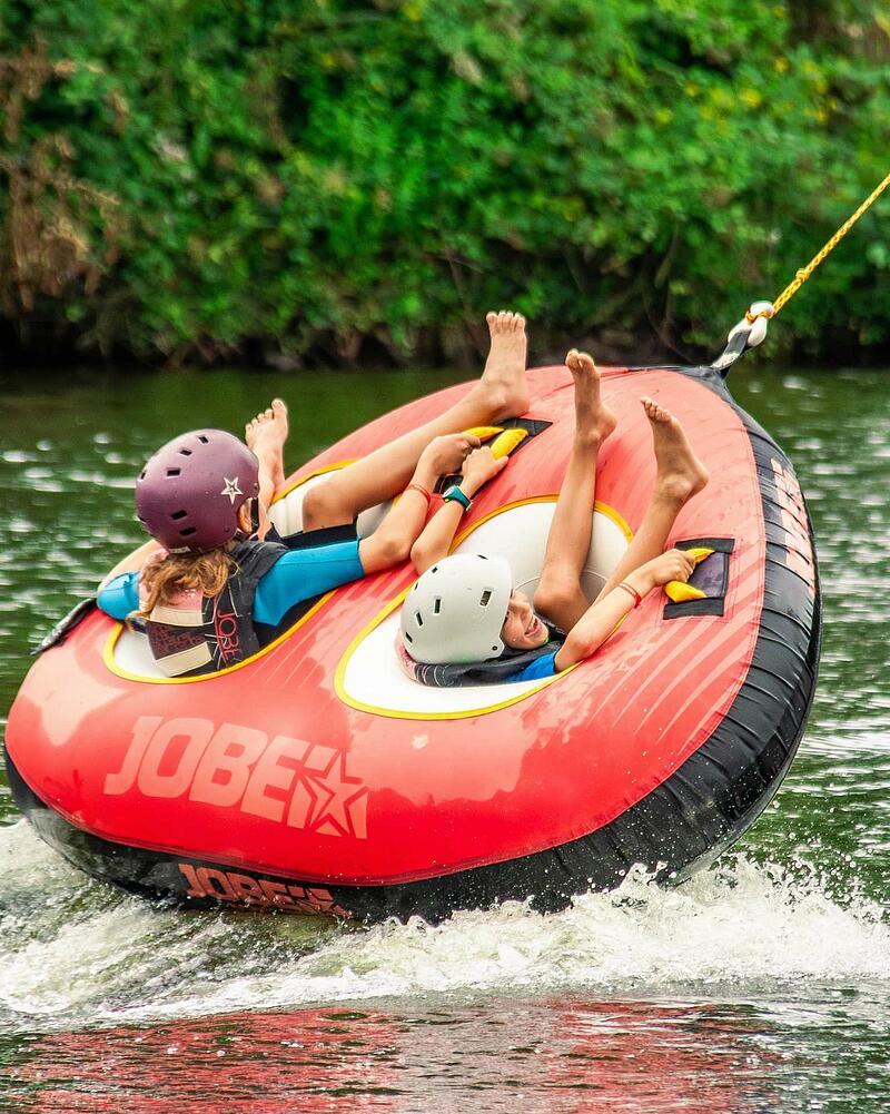 Two young kids wearing safety helmets and life vests scream with delight while riding a vibrant red inflatable towable tube on the water, illustrative of the exciting water activities at Les Castels seaside campings in France.