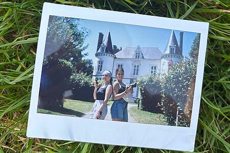 A Polaroid print resting on green grass, showing a photo of two young women with cameras posing back-to-back in front of a historic French castle.