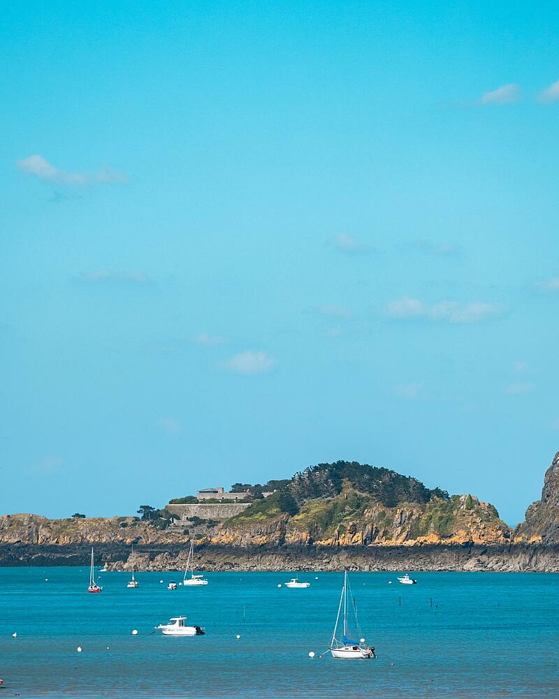 Sailboats and motorboats anchored on a turquoise blue sea facing a fortified rocky island under a clear blue sky in Brittany.