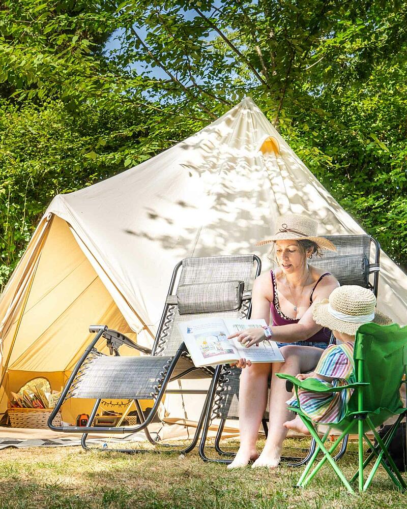 A mother and young child reading a book together in folding chairs outside a large canvas bell tent at a Les Castels 5-star campsite in France, representing premium campsites France.