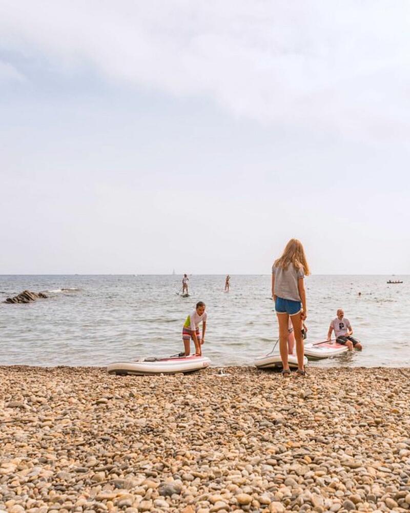 From a pebble beach in France, people prepare several stand-up paddleboards near the water's edge, illustrative of the relaxing water sports available to guests of Les Castels seaside campings.