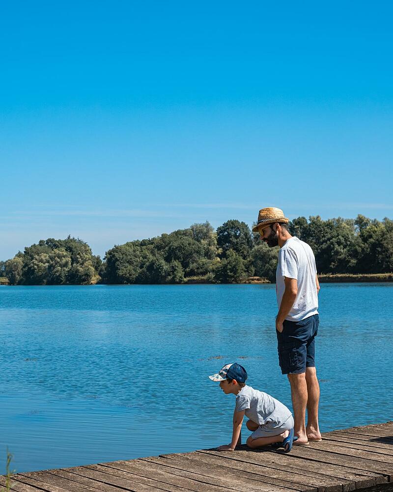 A father and son standing on a wooden pontoon looking out over a calm blue lake at a Les Castels 5-star campsite in France, showing the natural settings of premium campsites France.