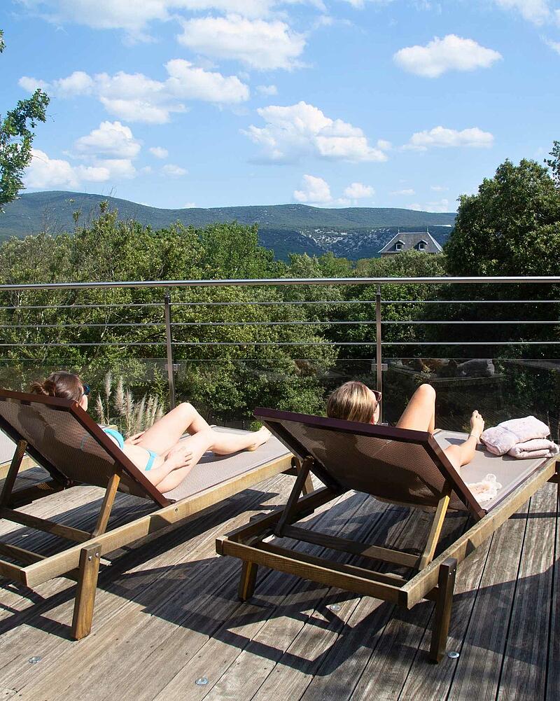 Two guests relaxing on sun loungers on a high wooden deck with panoramic mountain views at Les Castels Domaine de Sévenier campsite in Ardèche.