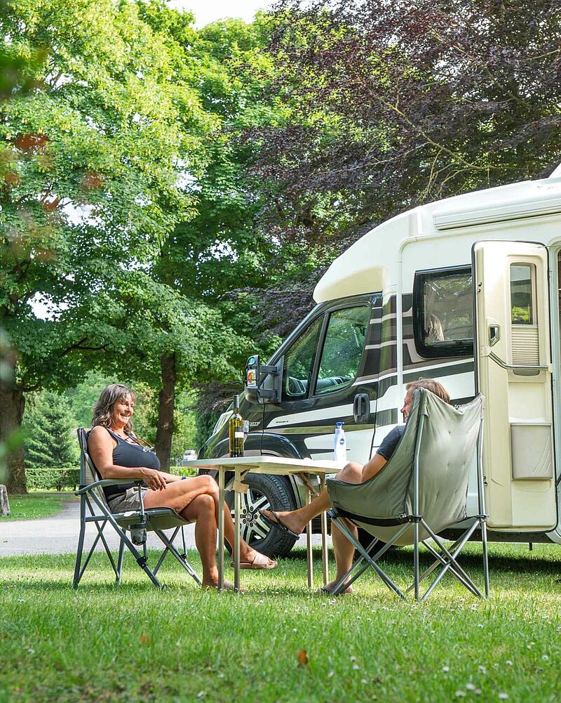 A man and a woman sit in comfortable folding chairs at a camping table with a bottle of wine, relaxing next to their modern motorhome on a shaded, grassy pitch.