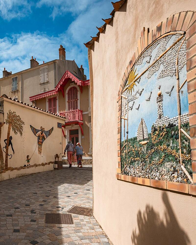 A charming European street in Île Penotte, close to a Les Castels campsite, featuring vibrant shell mosaic artwork on the walls of light-colored buildings.