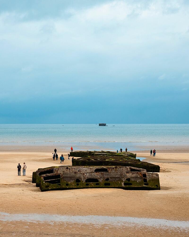 A panoramic view of a wide beach on the French coast, where tourists are exploring large, concrete military remains, which could be part of the D-Day Landing Beaches historical sites accessible from Les Castels seaside campings.