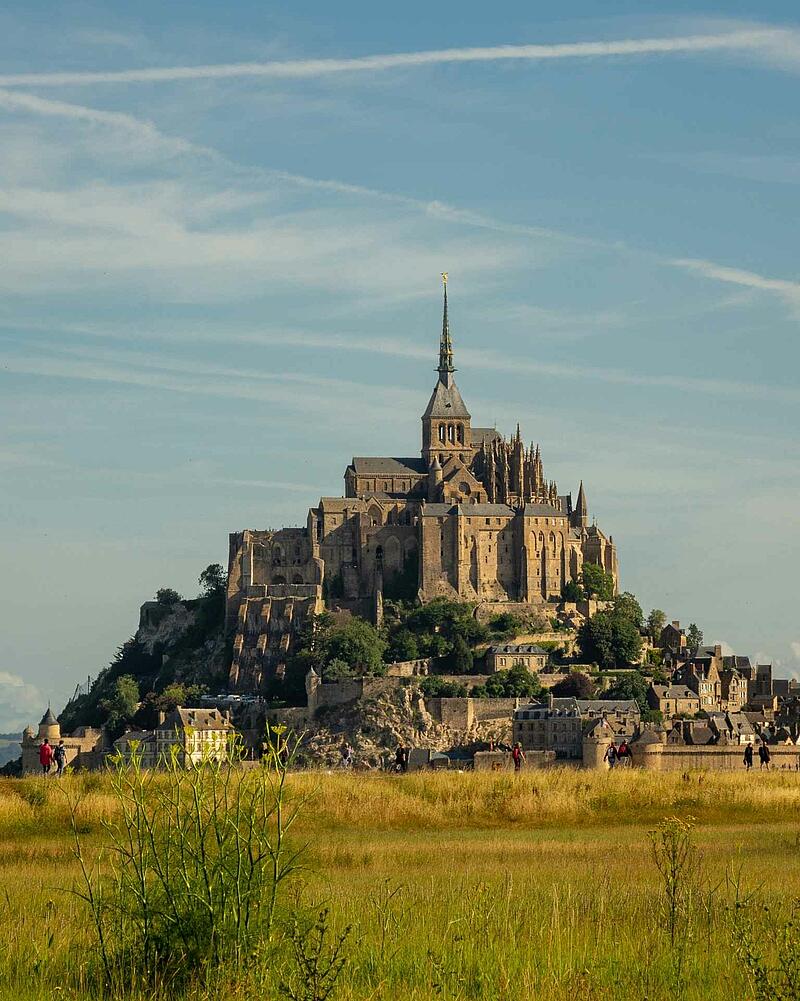 The magnificent tidal island of Mont-Saint-Michel with its towering medieval abbey, viewed from across a grassy green and yellow field under a light blue summer sky.