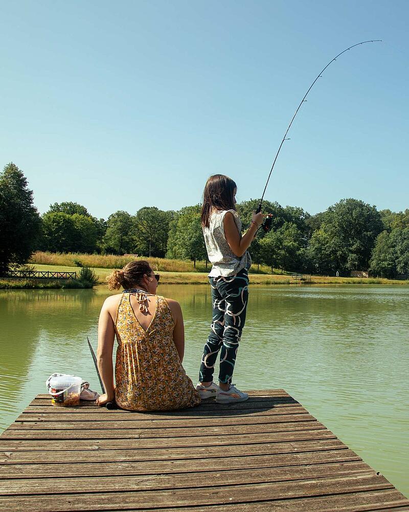 A mother and daughter are enjoying a peaceful day fishing together on a wooden pier by a serene lake, showcasing the relaxation and family-friendly activities during a last-minute outdoor holiday at Les Castels